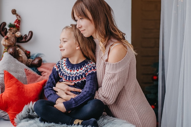 Dochter met moeder bij het raam wachtend op Kerstmis. Moeders tedere omhelzing. Aanhankelijke communicatie tussen moeder en dochter.