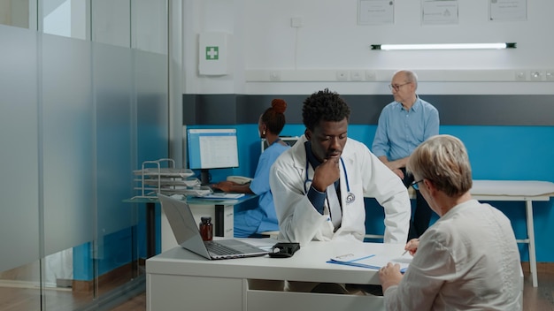 Doctor of african american ethnicity consulting old woman with sickness while sitting at desk in office. Senior patient signing checkup document files for healthcare appointment