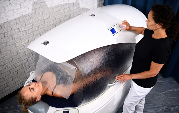 Doctor beautician adjusting programs in spa capsule with a woman inside it receiving professional beauty treatment