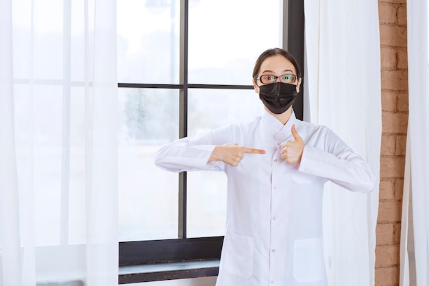 Doctor in black mask and eyeglasses standing by the window and showing enjoyment sign. 