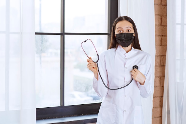 Doctor in black mask with a stethoscope standing by the window and checking the heartbeats. 