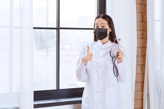 Doctor in black mask with a stethoscope standing by the window and showing satisfaction sign. 