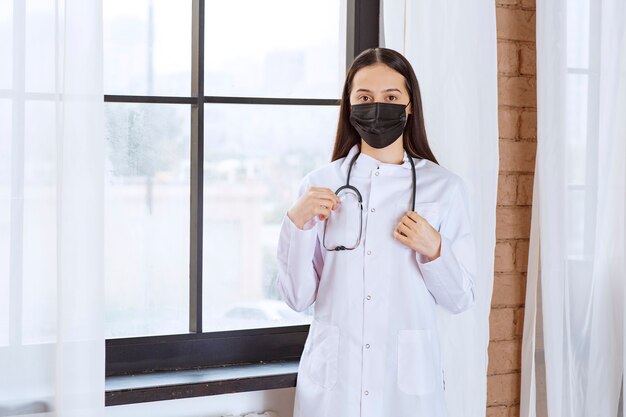 Doctor in black mask with a stethoscope standing by the window. 