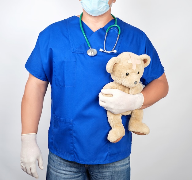 Doctor in blue uniform and white latex gloves holding a brown teddy bear