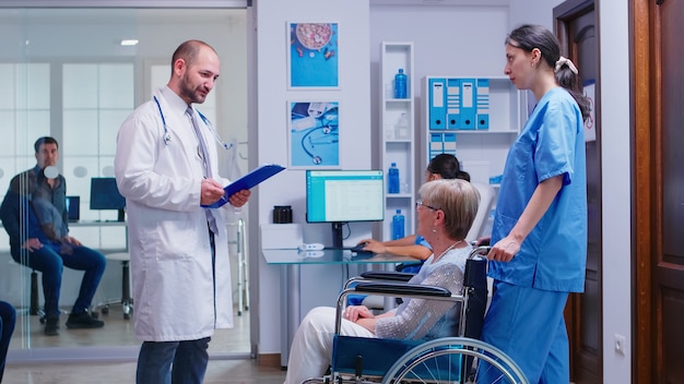 Doctor checking diagnosis of disabled senior woman in wheelchair. Medical staff in hospital hallway with invalid patient. Man in medical examination room.