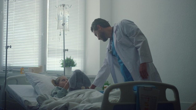 Doctor checking on little patient in hospital ward Sick girl hugging plush toy