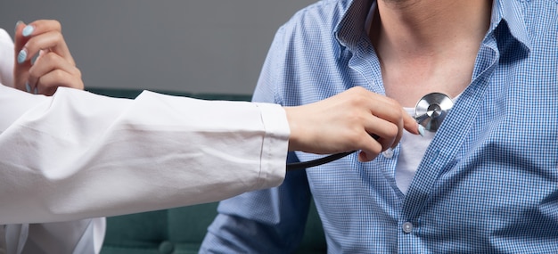 A doctor checks a man's heart with a stethoscope on the sofa