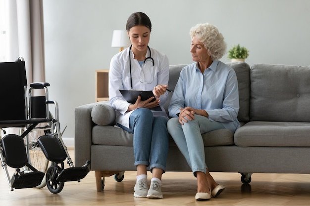 Doctor consulting disabled older woman filling medical form at clipboard writing prescription sitting on couch at home during visit physician wearing white uniform talking with mature