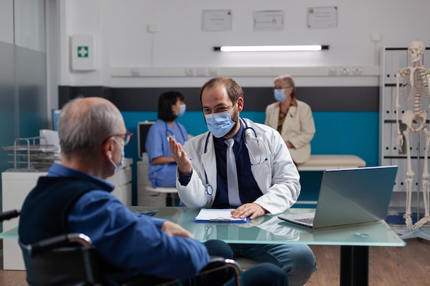 Doctor consulting elder man with physical disability in office, doing health care examination for recovery. Medic and patient sitting in wheelchair meeting during covid 19 pandemic.