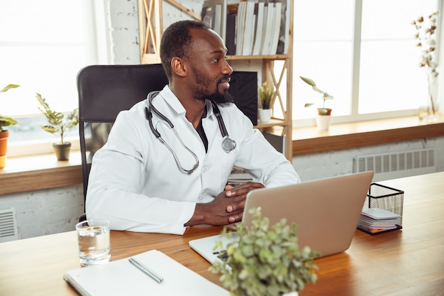 Consulenza medica per il paziente, lavorando con il computer portatile. medico afroamericano durante il suo lavoro con i pazienti, spiegando le ricette per la droga. duro lavoro quotidiano per la salute e il salvataggio di vite durante l'epidemia.