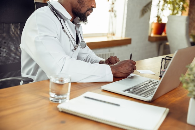 Doctor consulting for patient, working with laptop. African-american doctor during his work with patients, explaining recipes for drug. Daily hard work for health and lives saving during epidemic.