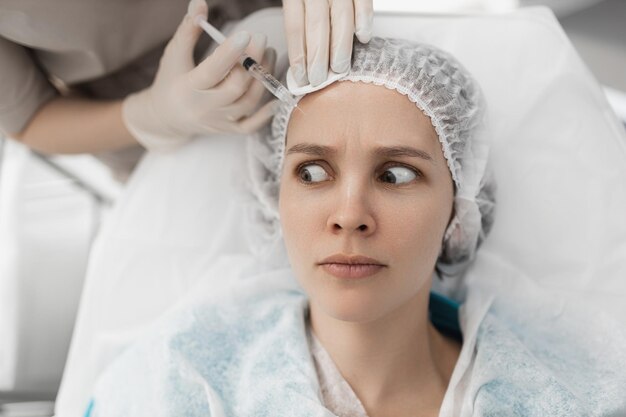 Doctor cosmetologist with a syringe in his hand makes injections in the forehead of the face of a frightened client