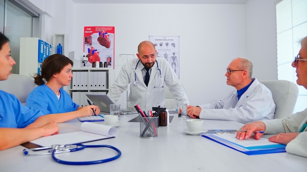 Doctor discussing with medical staff in hospital conference room wearing lab coat. Clinic expert therapist talking with colleagues about disease, diagnosis for patients treatment issues