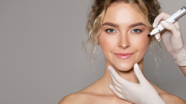 Photo doctor drawing marks on young womans face for cosmetic surgery operation over grey background