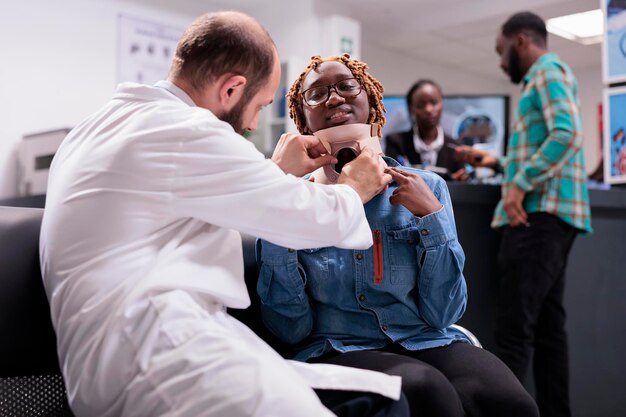 Doctor examining african american woman with neck collar, sitting in waiting room at hospital reception lobby. Patient injured in accident wearing cervical foam brace, healthcare recovery.