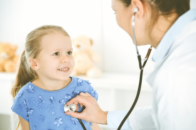 Doctor examining a child by stethoscope. Happy smiling girl patient dressed in blue dress is at usual medical inspection. Medicine concept.