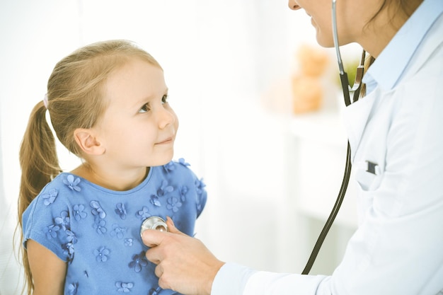 Врач осматривает ребенка стетоскопом. Счастливая улыбающаяся пациентка, одетая в синее платье, проходит обычный медицинский осмотр. Концепция медицины.