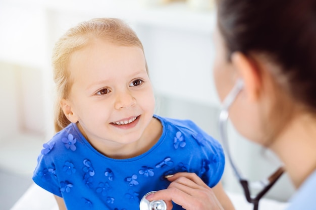 Врач осматривает ребенка с помощью стетоскопа в солнечной клинике. Счастливая улыбающаяся пациентка, одетая в синее платье, проходит обычный медицинский осмотр.
