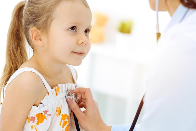 Doctor examining a child by stethoscope in sunny clinic. Happy smiling girl patient dressed in bright color dress is at usual medical inspection.