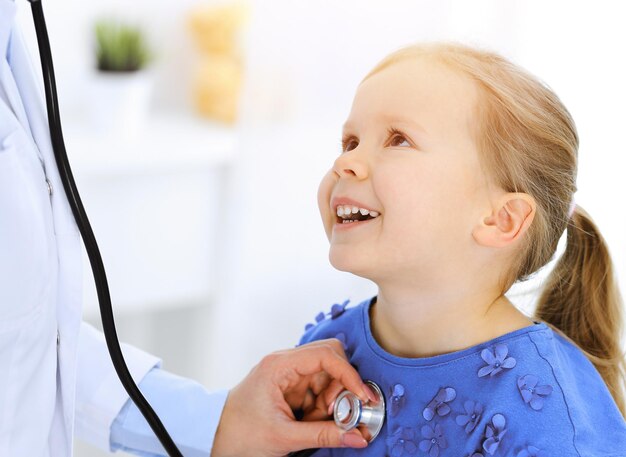 Doctor examining a little girl by stethoscope. Happy smiling child patient at usual medical inspection. Medicine and healthcare concepts.