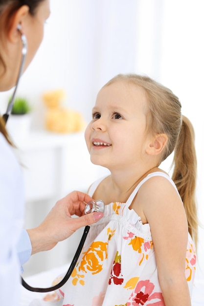 Doctor examining a little girl by stethoscope. Happy smiling child patient at usual medical inspection. Medicine and healthcare concepts.