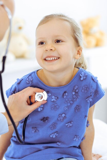 聴診器で少女を診察する医師。いつもの健康診断で幸せな笑顔の子供の患者。医学とヘルスケアの概念。