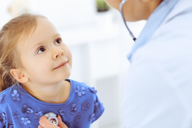 Doctor examining a little girl by stethoscope. Happy smiling child patient at usual medical inspection. Medicine and healthcare concepts.