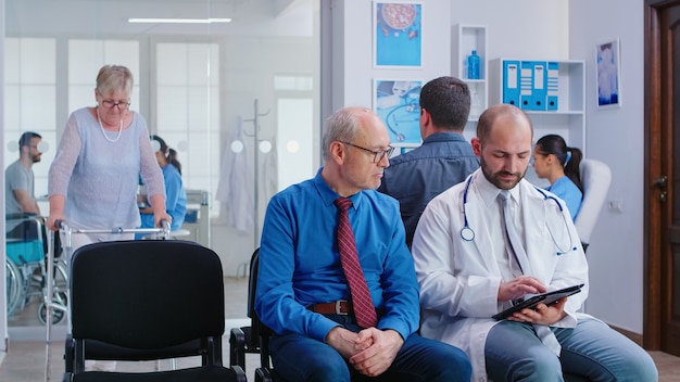 Doctor explaining test results to senior man in hospital waiting area. Disabled senior woman with walking frame arring for medical examination.