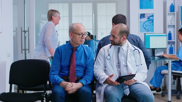 Doctor explaining treatment to senior man in hospital waiting area. Woman with walking frame arriving at medical examination.