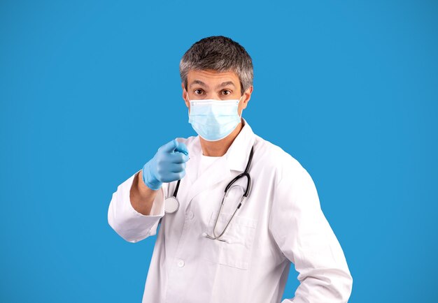 Doctor in facial mask pointing at camera over blue background