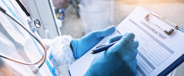 의사는 Covid19 코로나바이러스 전염병에서 창에 기대어 있는 진료소에서 보호복을 입은 펜으로 코로나바이러스 테스트 데이터 시트를 작성합니다