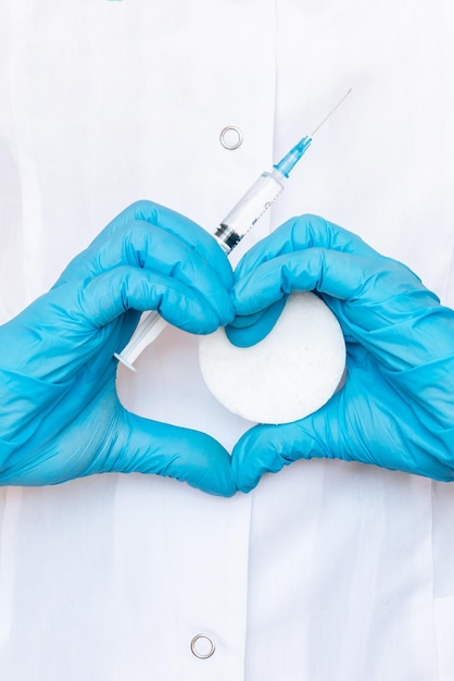 Foto il medico che forma una forma di cuore con le sue mani in guanti blu che tengono una siringa e un batuffolo di cotone