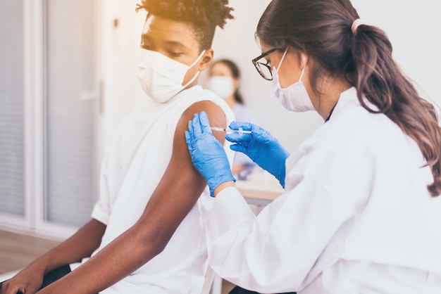 Doctor give a shot to african boy wearing face mask getting vaccinated during coronavirus or covid-19 pandemic at hospital,Vaccination concept