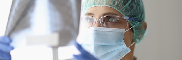 Doctor in gloves and protective mask examines X-ray of lungs with pneumonia of patient with covid19.