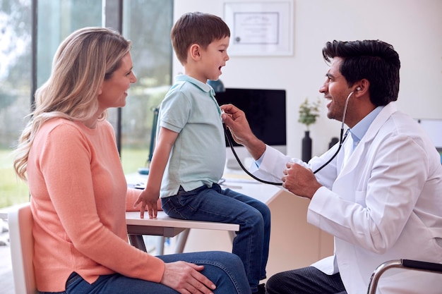 Doctor Or GP In White Coat Meeting Mother And Son For Appointment In Office Listening To Chest