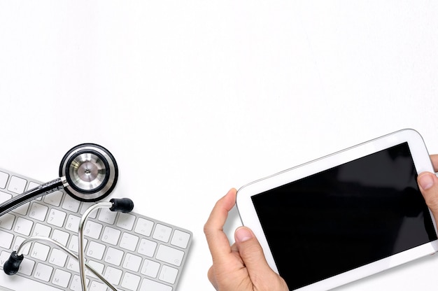 白いテーブルに白いキーボードに空白の画面と聴診器を持つ医師の手持ち株タブレット