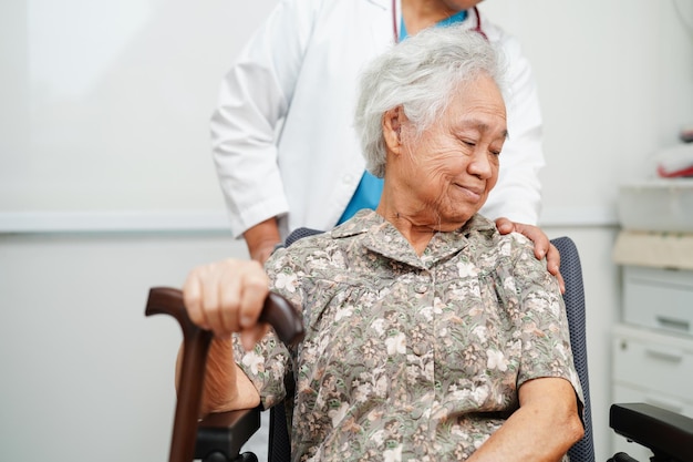 Doctor help Asian elderly disability woman patient holding walking stick in wrinkled hand at hospital