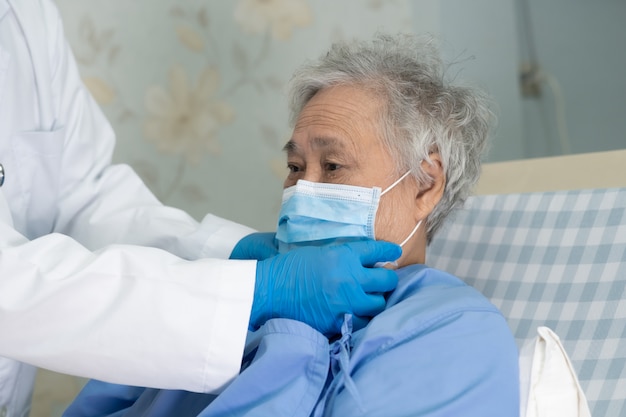 의사는 병원에서 얼굴 마스크를 쓰고 아시아 수석 여자 환자가 Covid-19 바이러스를 보호하도록 도와줍니다.