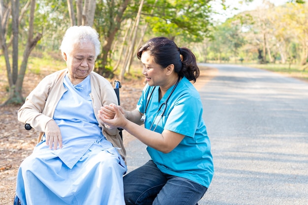 医者は公園で車椅子に座っているアジアの年配の女性患者を助け、世話をします。