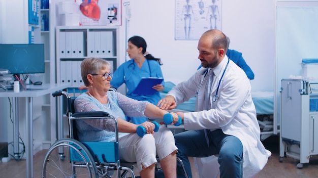 Doctor helping disabled senior patient to recover muscle strength in private modern rehabilitation clinic or hospital. nurse in background talking with elderly man with walking frame