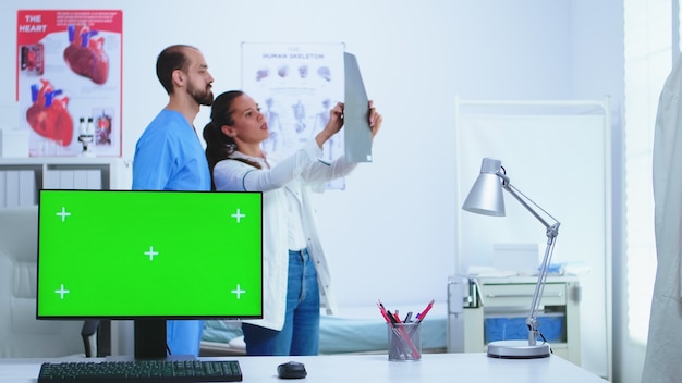 Foto medico e il suo assistente che controllano i raggi x del paziente e il computer con display verde in ospedale. desktop con schermo sostituibile in clinica medica mentre il medico controlla la radiografia del paziente per la diagnosi