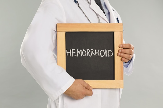 Doctor holding blackboard with word HEMORRHOID on light grey background closeup