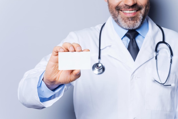 Doctor holding copy space. Close-up of confident mature doctor showing his business card and smiling while standing against grey background