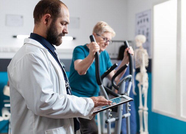 Doctor holding digital tablet for checkup with woman at\
recovery. senior patient using stationary bicycle for physical\
exercise while talking to medical specialist about\
physiotherapy