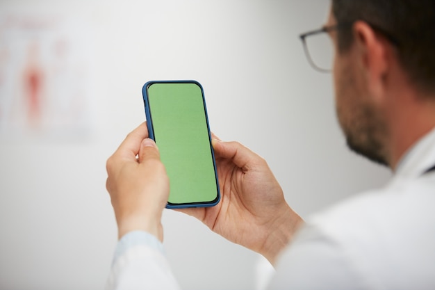 Doctor holding green screen smartphone, senior physician using chroma key phone sitting in medical cabinet in white gown and gasses