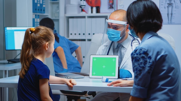 Doctor holding green screen tablet in medical office sitting on the desk. Healthcare specialist with chroma key notebook isolated mockup replacement screen. Easy keying medicine medical related theme.