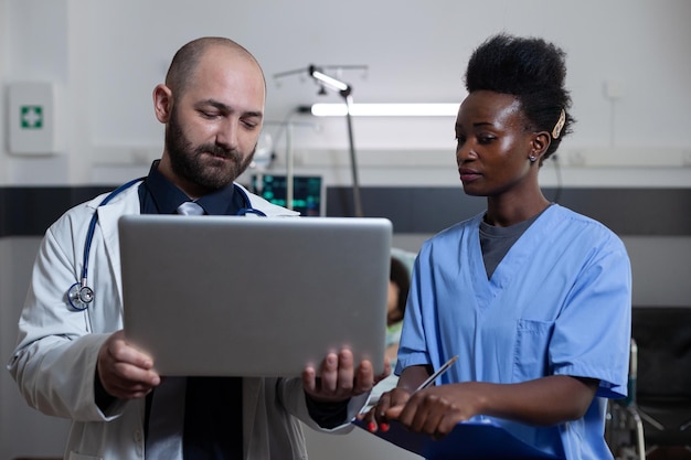 Doctor holding laptop presents patient medical history to nurse holding clipboard with lab results in hospital ward. Medical specialist discussing digital examination report with health care worker.