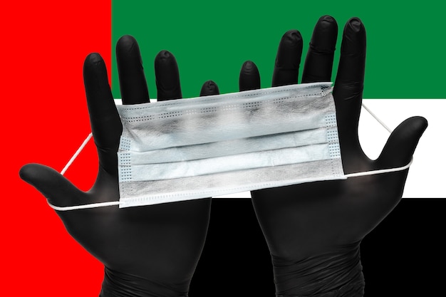 Doctor holding medical mask for face in two hands black gloves on background national flag of united arab emirates uae. concept quarantine, pandemic insurance coronavirus, outbreak airborne diseases.