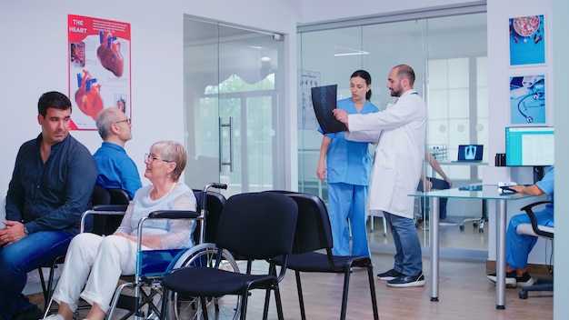 Doctor holding patient x-ray explaining diagnosis to nurse in waiting area. Disabled senior woman wheelchair waiting for medical examination. Hospital and health care system, medicine private clinic h