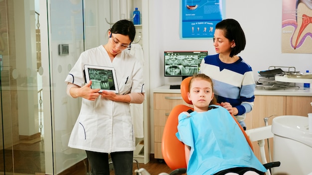 간호사가 백그라운드에서 도구를 준비하는 동안 의사가 엑스레이로 태블릿을 들고 여자 환자의 어머니에게 태블릿을 보여줍니다. 구강 클리닉에서 현대적인 장치를 사용하여 치과 방사선 촬영을 제시하는 구강 전문의